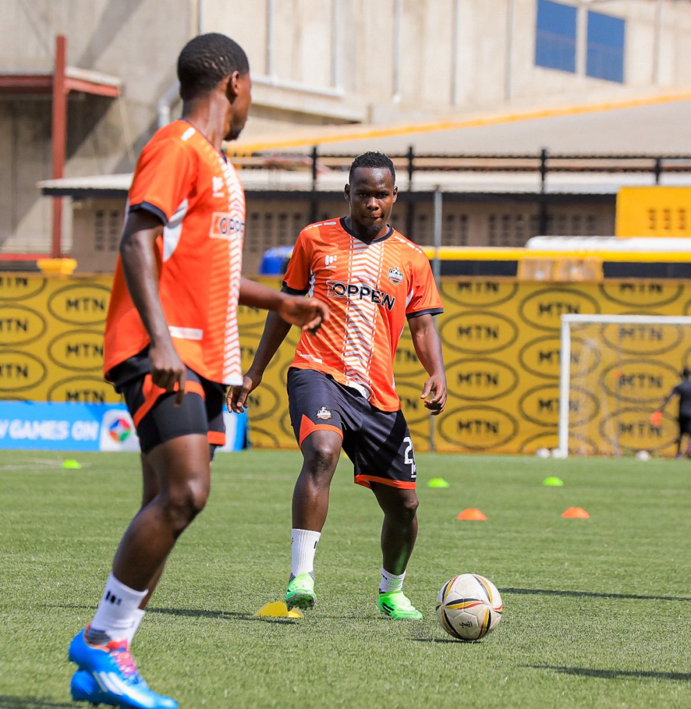 Two NEC FC players in orange training kit work with the ball on the pitch during a training session, with cone markers set out and MTN branded boards in the background.