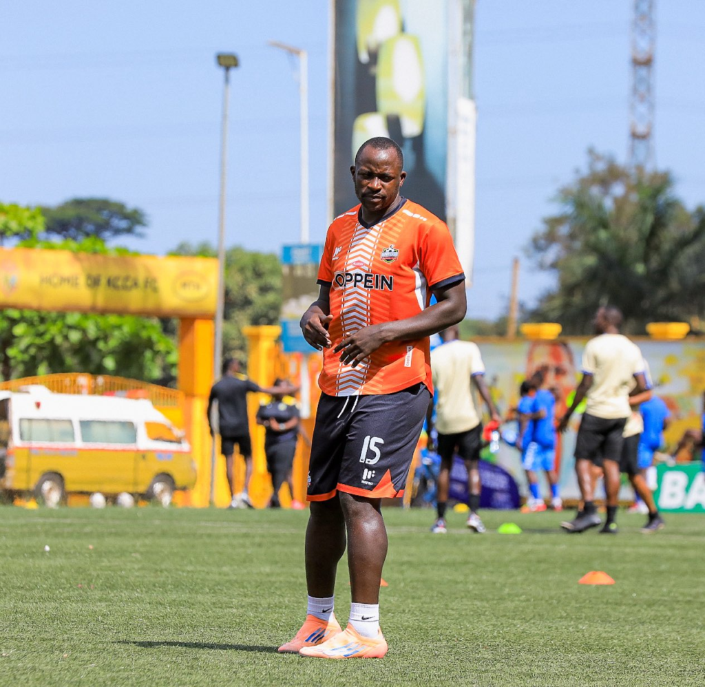 An NEC FC player in an orange OPPEIN-branded jersey, number 15, stretches on the pitch during a warm-up session, with other players and a stadium gate visible in the background.