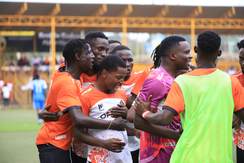 A group of NEC FC players in orange, white, and pink kits laugh and embrace in a close huddle while celebrating a goal on the pitch.