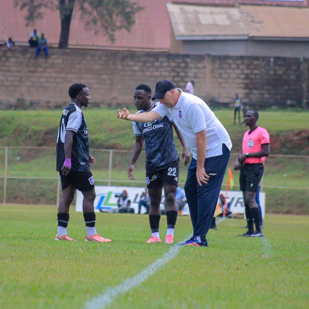 SC Villa coach Zeljko Kovacevic, wearing a cap and white polo shirt, gestures while speaking to player Patrick Kakande on the sideline as a teammate and referee look on.