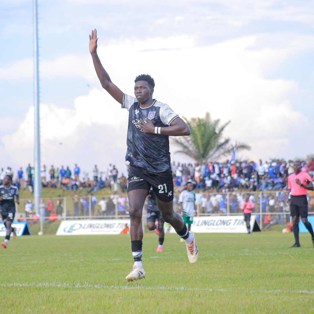 SC Villa player Otim Andrew, number 21, celebrates after scoring against Kigezi Home Boys, raising his hand to a packed stand behind him.
