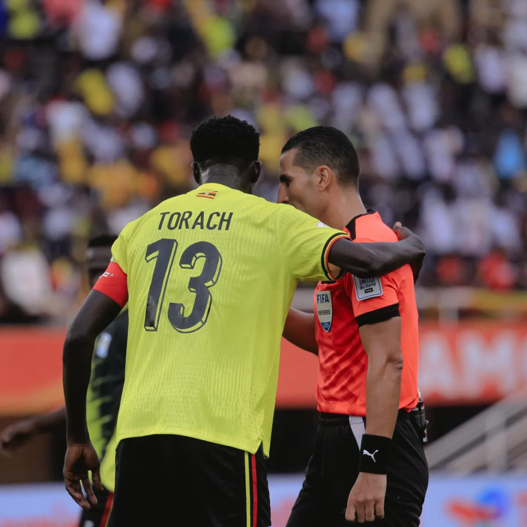 Rogers Torach Ochaki, wearing Uganda’s number 13 shirt and captain’s armband, communicates with a FIFA referee on the pitch.