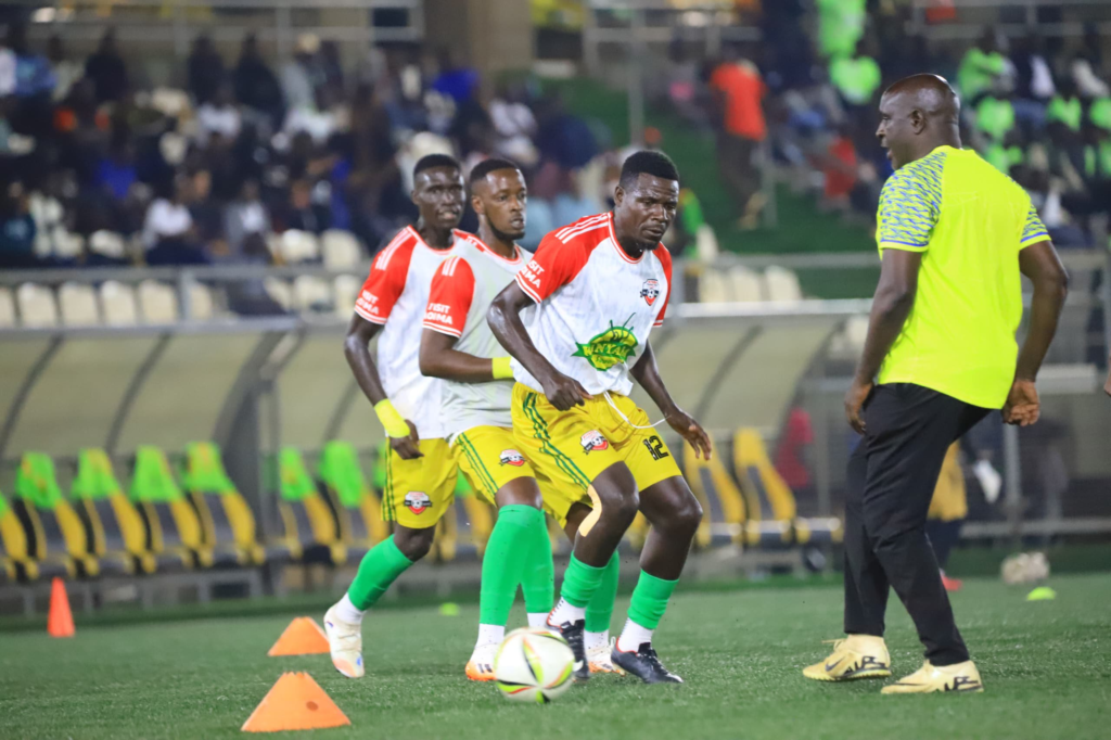 Kitara FC players in white, red, yellow, and green training kits take part in a warm-up drill under a coach’s supervision.