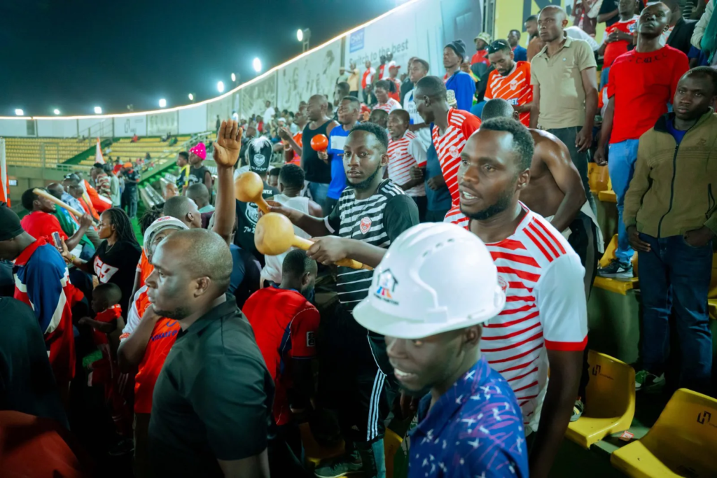 Express FC fans wearing red-and-white club colours cheer and play instruments in the stands during a match at Hamz Stadium.