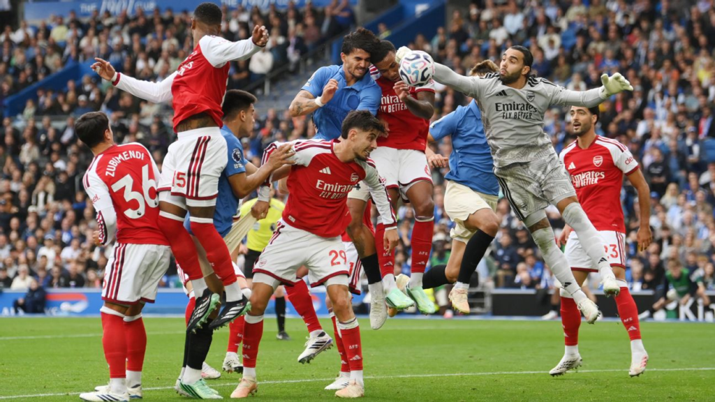 Arsenal goalkeeper punching the ball away from a crowded penalty area during an aerial challenge with players from both teams.