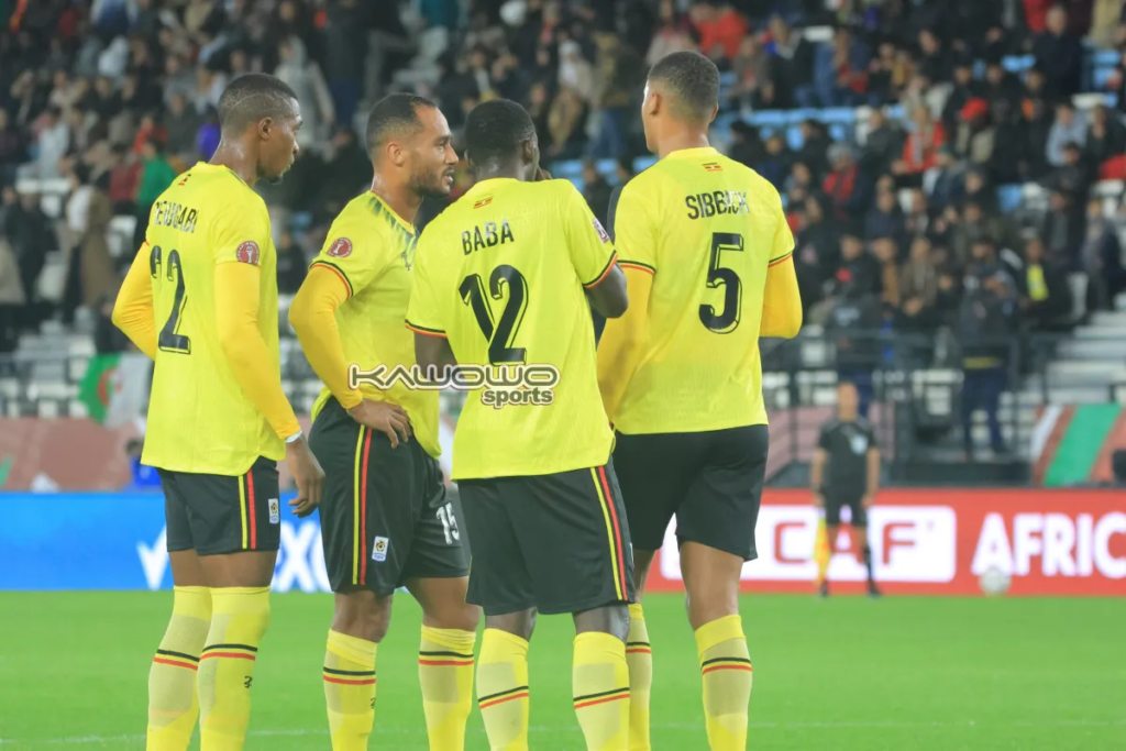 Four Uganda Cranes players in yellow kits huddled together on the pitch, mostly facing away from the camera, with Baba (No. 12) and Sibbick (No. 5) visible by their shirt names, and spectators in the stands behind them.