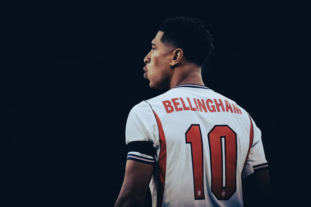 England player Jude Bellingham, wearing the captain's armband and No. 10 shirt, pictured from behind against a dark background.