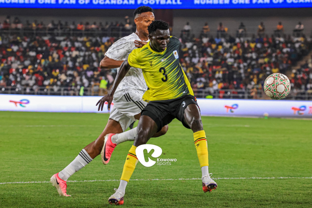 Uganda Cranes player Rogers Torach, wearing No. 3, shielding the ball from an opponent in white as it goes out of play, in front of a packed stadium.