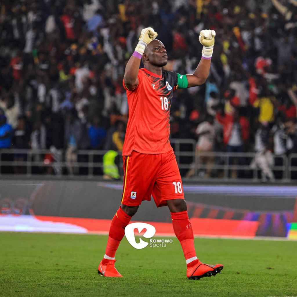 Uganda Cranes goalkeeper Denis Onyango, wearing No. 18 and gloves, raising both fists in celebration in front of a packed stadium.
