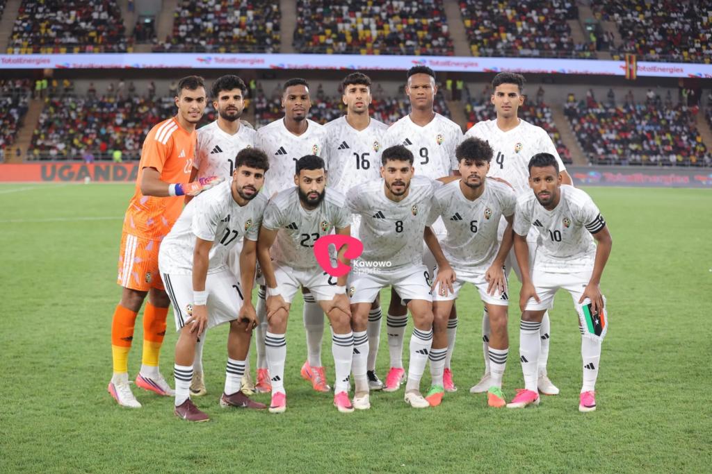 The Libya starting XI posing for a team photo in white kits before their match against Uganda, with a packed stadium in the background.