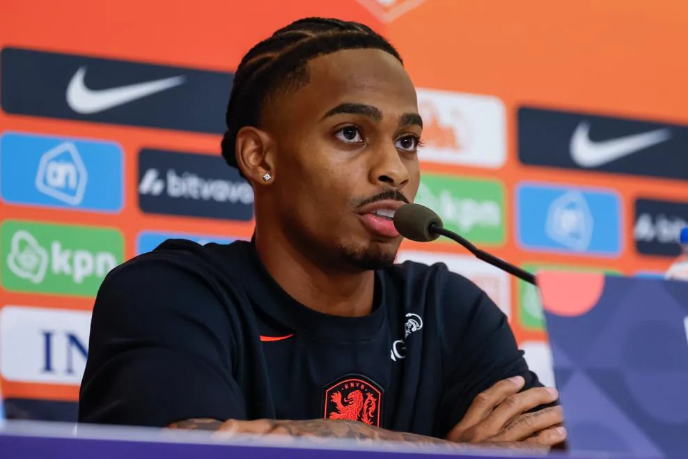 Netherlands forward Crysencio Summerville speaking into a microphone at a press conference, wearing a black Netherlands training top.