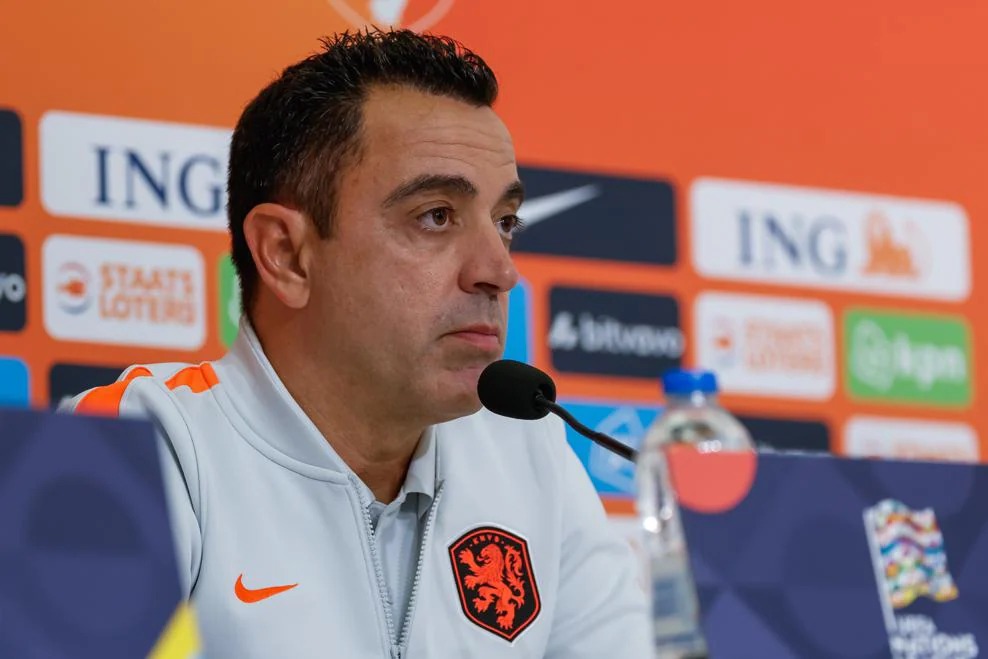 Netherlands head coach Xavi Hernández speaking into a microphone at a press conference, wearing a light grey Netherlands training top.