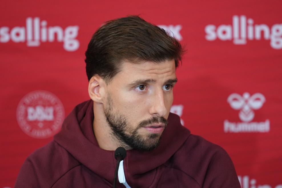 Portugal defender Rúben Dias, wearing a maroon hoodie, speaking at a press conference in front of a red backdrop with Danish Football Association (DBU) and hummel branding.