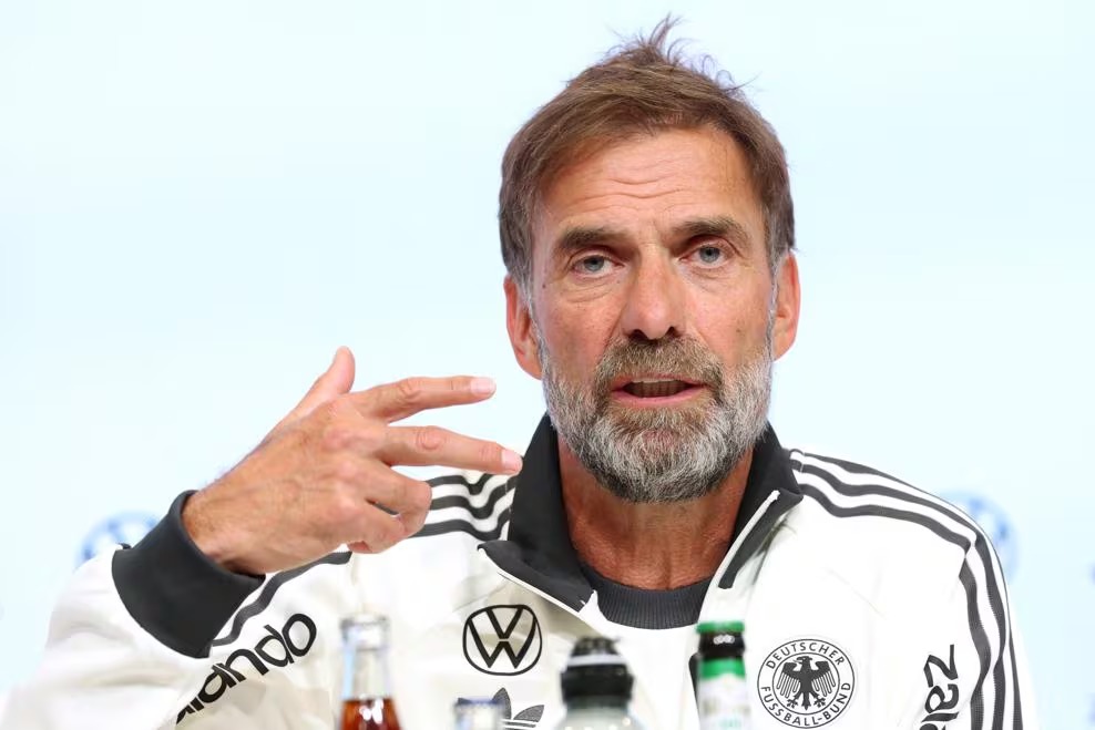 Germany head coach Jürgen Klopp gesturing while speaking at a press conference, wearing a white Germany tracksuit top.