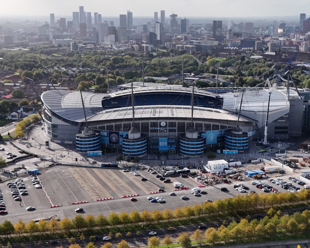 Aerial drone view of Manchester City's empty Etihad Stadium, showing the blue seating bowl and pitch from above.