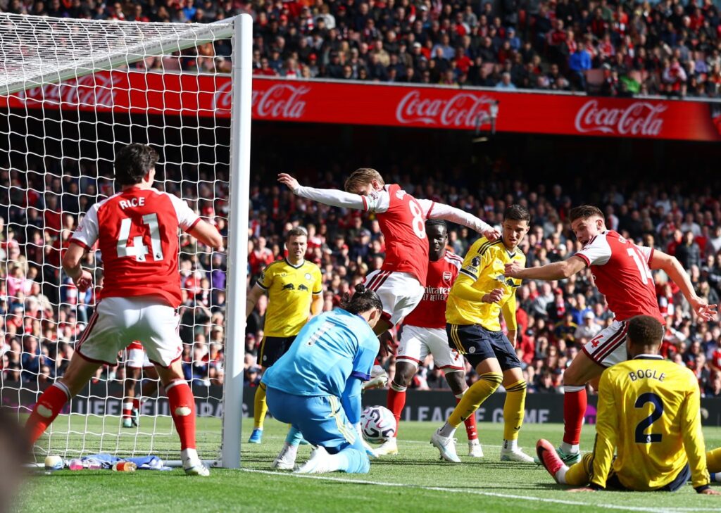 Leeds goalkeeper James Trafford, in light blue wearing No. 1, gathering the ball on the ground by the post as Arsenal’s Martin Odegaard (No. 8) leans over him. Arsenal’s Rice (No. 41), a teammate in No. 14 and Leeds’ Bogle (No. 2, seated) are around the goalmouth, in front of a packed Emirates crowd.