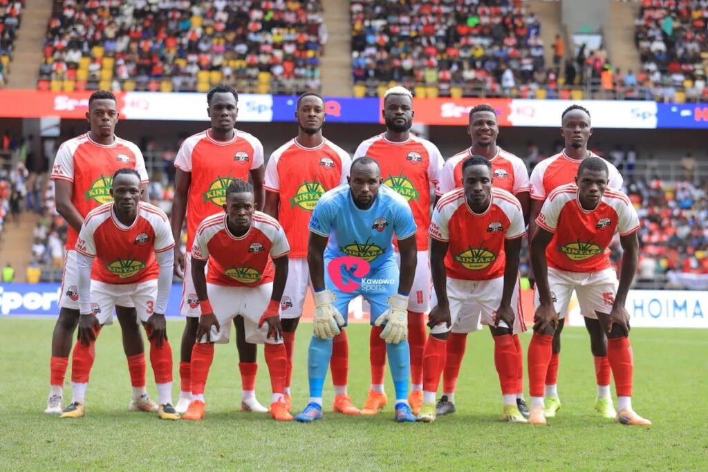 The Kitara starting XI posing for a team photo before their match against Al Ahly, six players standing at the back and five crouching in front, in red and white Kinyara Sugar kits. The goalkeeper, in the middle of the front row, wears light blue with gloves, and the stands behind them are packed.