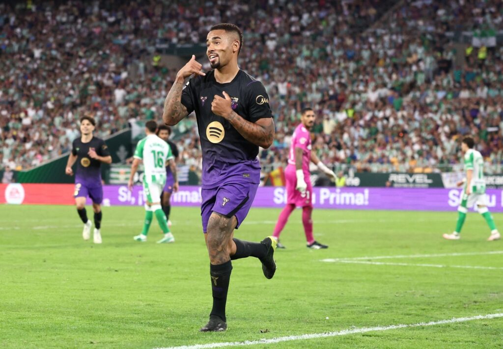 Barcelona’s Gabriel Jesus, in a black and purple kit, runs across the pitch smiling, with one hand held to his ear like a phone and the other on his chest. Behind him, a teammate in the same kit runs in from the left, with players in green and white shirts, a goalkeeper in pink and a packed stand in the background.