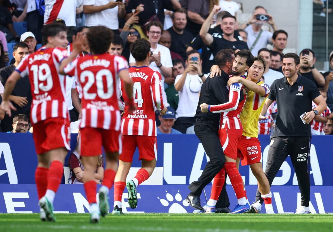 Atlético Madrid’s Álex Baena, in a red and white striped kit, is held in a hug by a man in black with his back to the camera, while a teammate in a yellow training bib has an arm around him. Teammates wearing No. 19, No. 20 and No. 4 run toward them from the left, and a staff member in black on the right smiles. Fans fill the stand behind, many cheering and holding up phones, in front of a blue “LaLiga” advertising board.