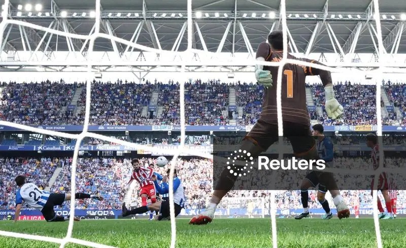 Seen from behind the goal net, a goalkeeper wearing No. 1 in a brown kit and green gloves stretches sideways as the ball flies past him. Atlético Madrid’s Alex Baena, in red and white stripes, stands in the middle of the penalty area with his arms out, and Espanyol defenders in blue and white are around him, one stretched out on the grass. A full stand rises behind them.