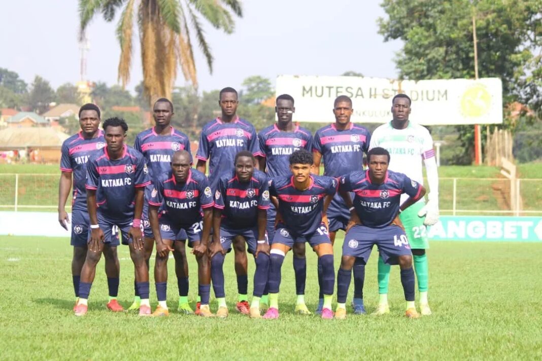 Kataka FC’s starting eleven pose for a team photo on the pitch in two rows. Ten players wear navy shirts with pink trim and “Kataka FC” on the chest, and the goalkeeper, standing at the back right, wears a white and green kit with green gloves. The front row crouches and the back row stands. A “Mutesa II Stadium” sign, a palm tree and a fence are behind them.