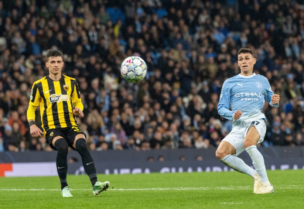 Manchester City’s Phil Foden, wearing No. 47 in a sky blue kit, follows through after striking the ball with his fists clenched and his eyes on it. An opponent in a yellow and black striped kit stands to the left, also watching the ball as it flies in the air between them. A packed stand and a Champions League advertising board are behind them.