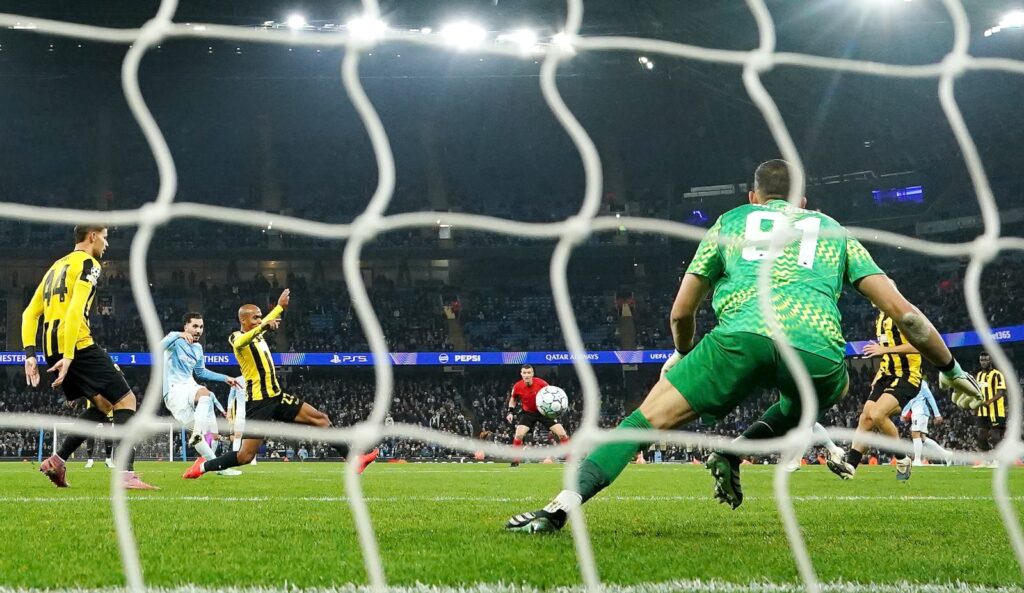 Seen from behind the goal net, Manchester City’s Rayan Cherki, in a sky blue kit, shoots from the edge of the penalty area as a defender in a yellow and black kit stretches a leg out to block. Another defender, wearing No. 44, stands at the left. The goalkeeper in a green kit with No. 91 crouches in front of the net, and a referee in red stands in the middle of the pitch with the ball just beside him. A floodlit stadium fills the background