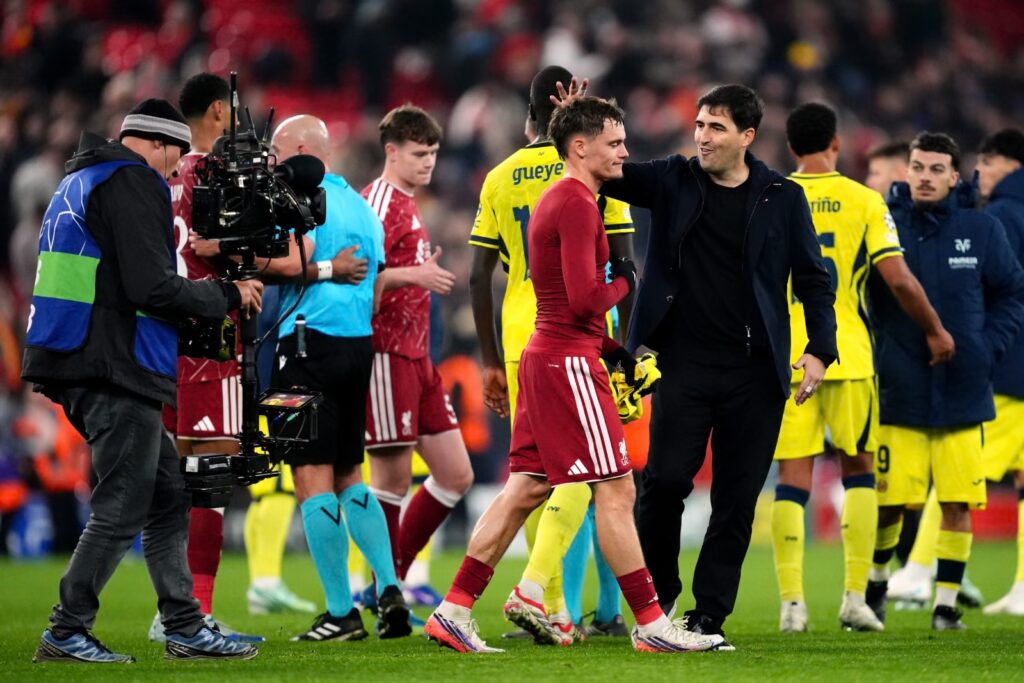 Liverpool manager Andoni Iraola, in a dark jacket and trousers, smiles and rests a hand on the shoulder of Liverpool’s Florian Wirtz, who walks past in a red kit with his head down, holding a pair of yellow gloves. A cameraman with a TV camera on the left, a referee in a light blue shirt, and Liverpool and Villarreal players in red and yellow are on the pitch around them.