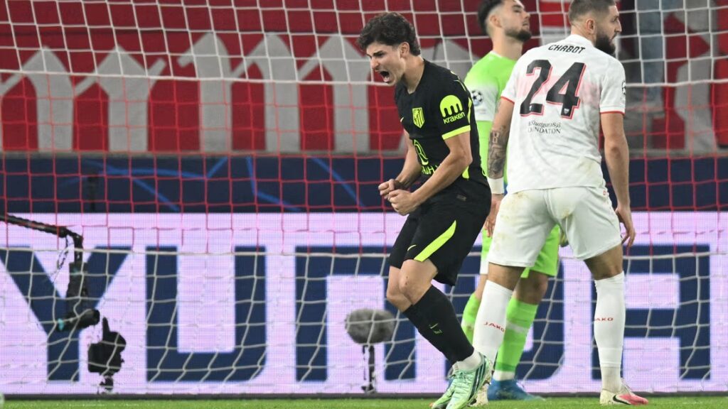 Atlético Madrid’s Julian Álvarez, in a black kit with neon green trim and the Kraken logo, clenches his fists and shouts with his mouth wide open. A Stuttgart player in white, wearing No. 24 with “Chabot” on the back, stands with his back to the camera, and a goalkeeper in a green kit is behind them. The goal net hangs in front of the camera, with a lit advertising board and a red banner behind.