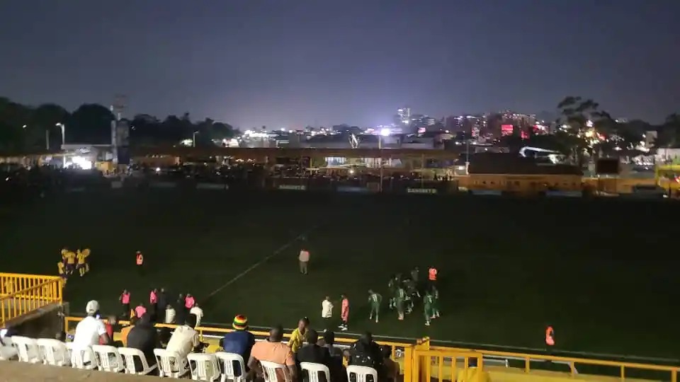 A night view across the pitch at MTN Omondi Stadium, which is almost entirely dark apart from a faint glow on the grass. A small group of players in green kits stand clustered near the centre, a few people in orange bibs and others in pink and white stand nearby, and a cluster of players in yellow stand at the far left. Spectators sit on white plastic seats in the foreground behind a yellow railing, with the lit buildings of the city skyline in the distance.