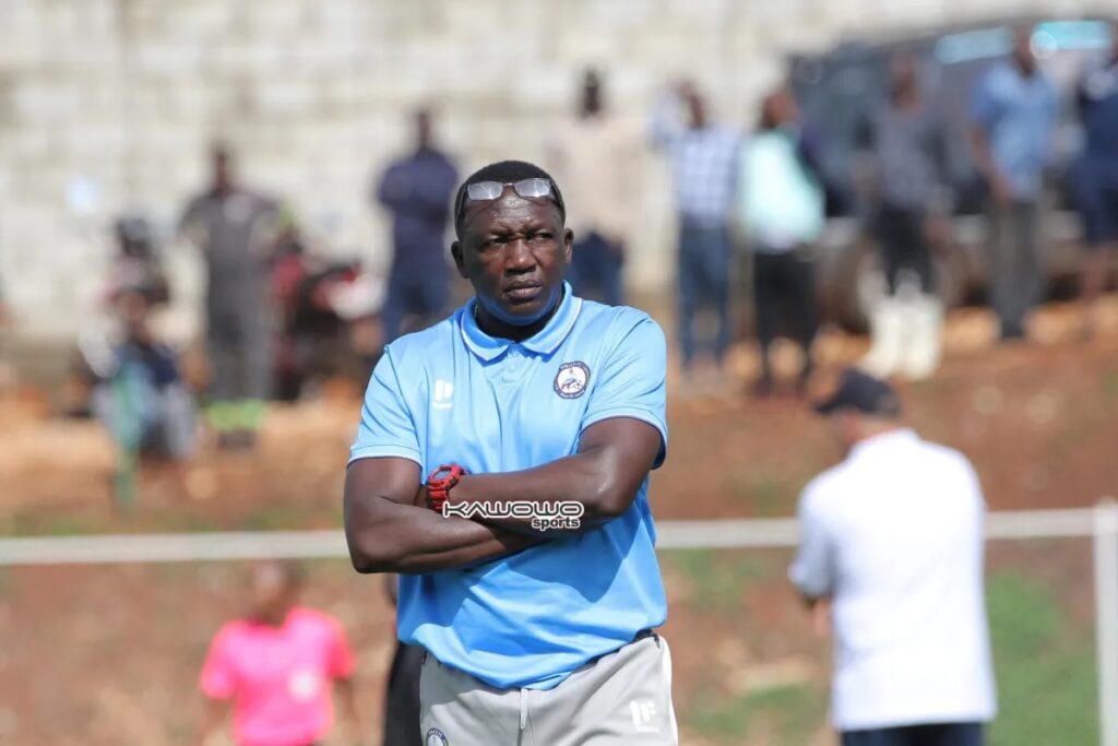 Police FC head coach Matia Lule, in a light blue polo shirt with the club crest, stands with his arms folded and a red watch on his wrist, glasses resting on his head, frowning. Blurred spectators stand on a red-brown bank behind him, with a man in white and a person in pink on the touchline.