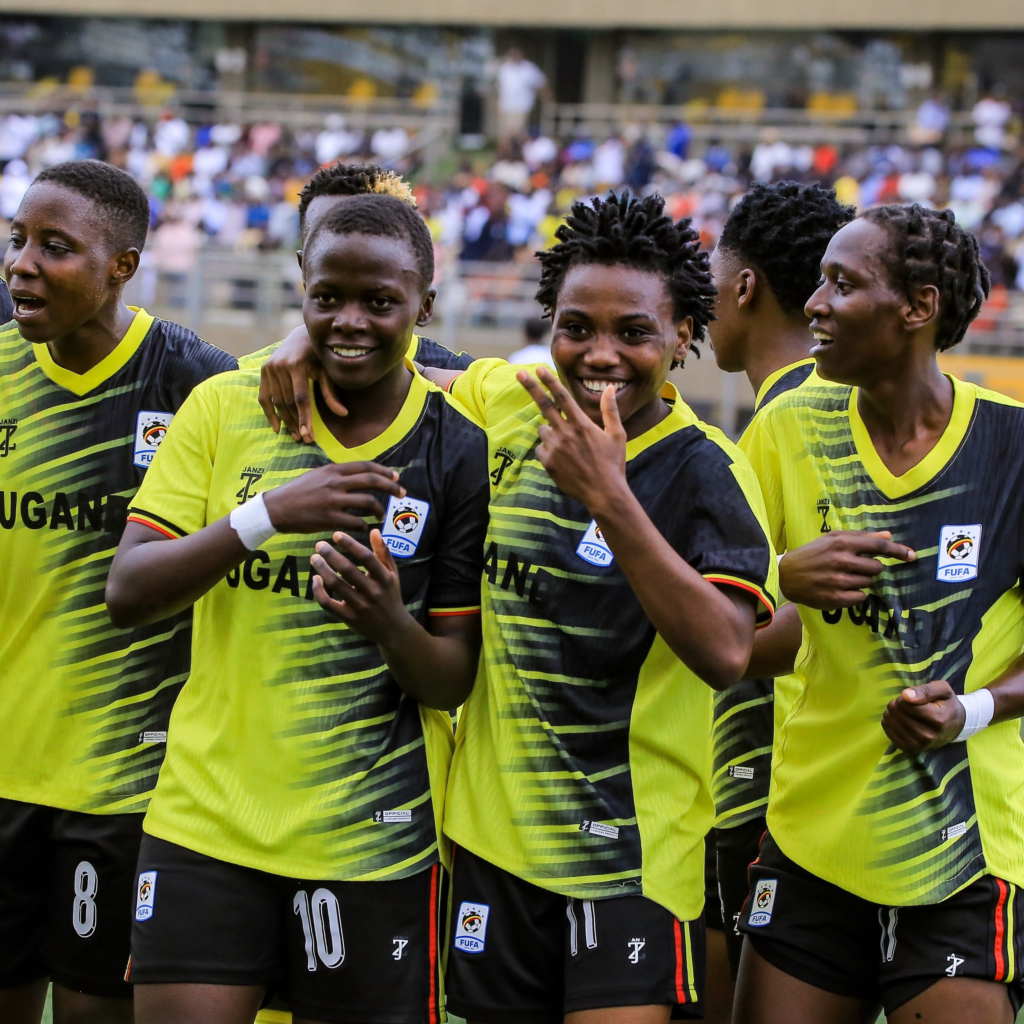 Uganda Crested Cranes players in yellow and black kits smiling and celebrating together on the pitch in front of a crowded stand, following Sylvia Kabene's goal.
