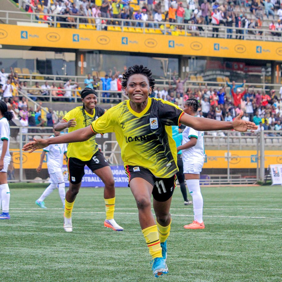 Uganda Crested Cranes forward Sylvia Kabene, wearing No. 11 in a yellow and black kit, smiling with arms spread wide as she celebrates a goal, with a teammate and a crowded stand behind her.