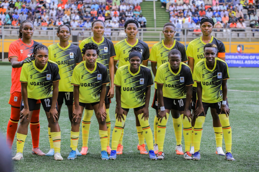 The Uganda Crested Cranes starting XI posing for a team photo in yellow and black kits ahead of their match against Zambia, with the goalkeeper in red at the back left and the captain in an orange armband at the back right, in front of a crowded stand.