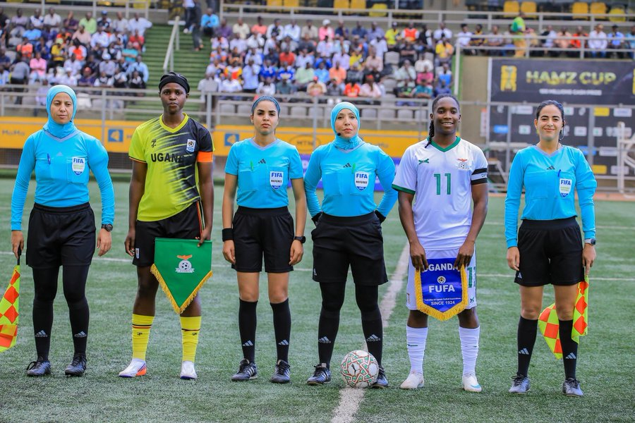 The Uganda and Zambia captains posing with four female match officials in light blue kits before kickoff, each captain holding the other team's pennant, with a Hamz Cup banner and crowded stand behind them.
