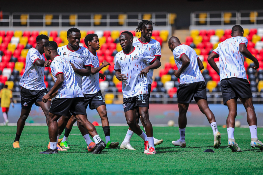 Vipers SC players in white kits with red, blue and black pinstripes warming up on the pitch ahead of their match against Black Powers, in front of an empty red, yellow and black stand.