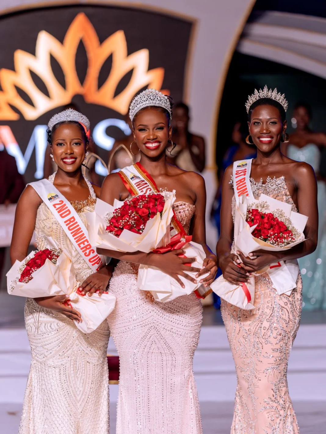 Three women in beaded gowns and tiaras holding bouquets of red roses on stage. The woman in the centre wears a Miss Uganda sash, with the 2nd runner-up on the left and the 1st runner-up on the right, in front of a Miss Uganda logo.