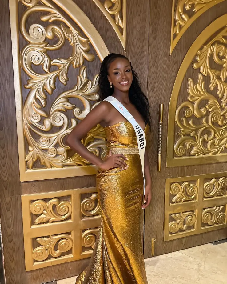 Almorie Sophia Nagayi, Miss Uganda 2026, smiling in a strapless gold gown and a white “Uganda” sash, posing with one hand on her hip in front of ornate gold-and-wood doors.