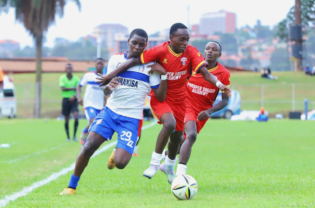 A SMAKISA player in a white and blue kit, wearing No. 29, shoulder to shoulder with two Prosper Soccer Academy players in red as they chase the ball on a grass pitch, with a referee and city buildings in the background.
