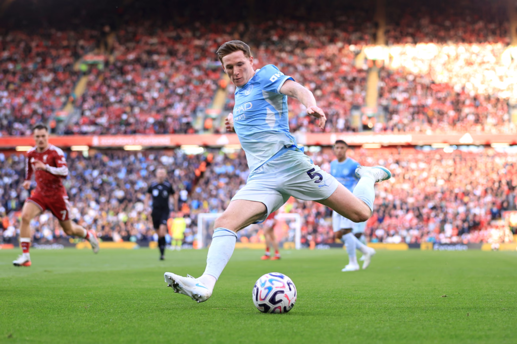 Manchester City’s Anderson, wearing a light blue No. 5 kit, stretching to strike the ball near the touchline, with a Liverpool player in red behind him and a packed Anfield crowd in the background.