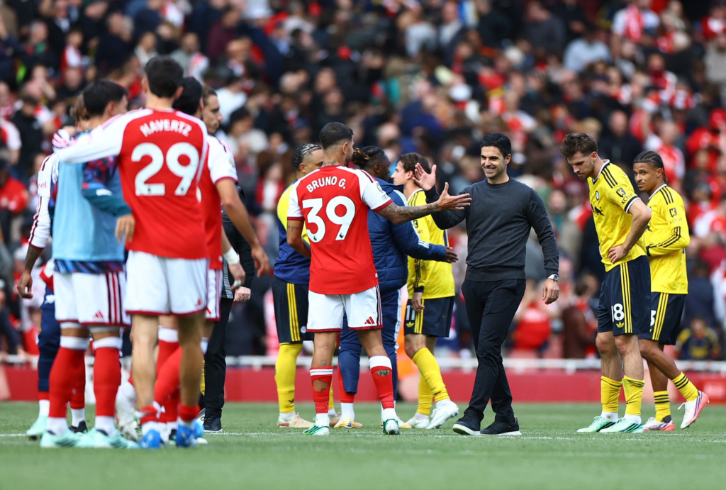 Arsenal manager Mikel Arteta smiling and shaking hands with Bruno Guimaraes, wearing No. 39 with “Bruno G.” on the back of his shirt, on the pitch after the final whistle. Dejected opponents in yellow stand to the right, while Arsenal players, including Havertz in No. 29, gather on the left.