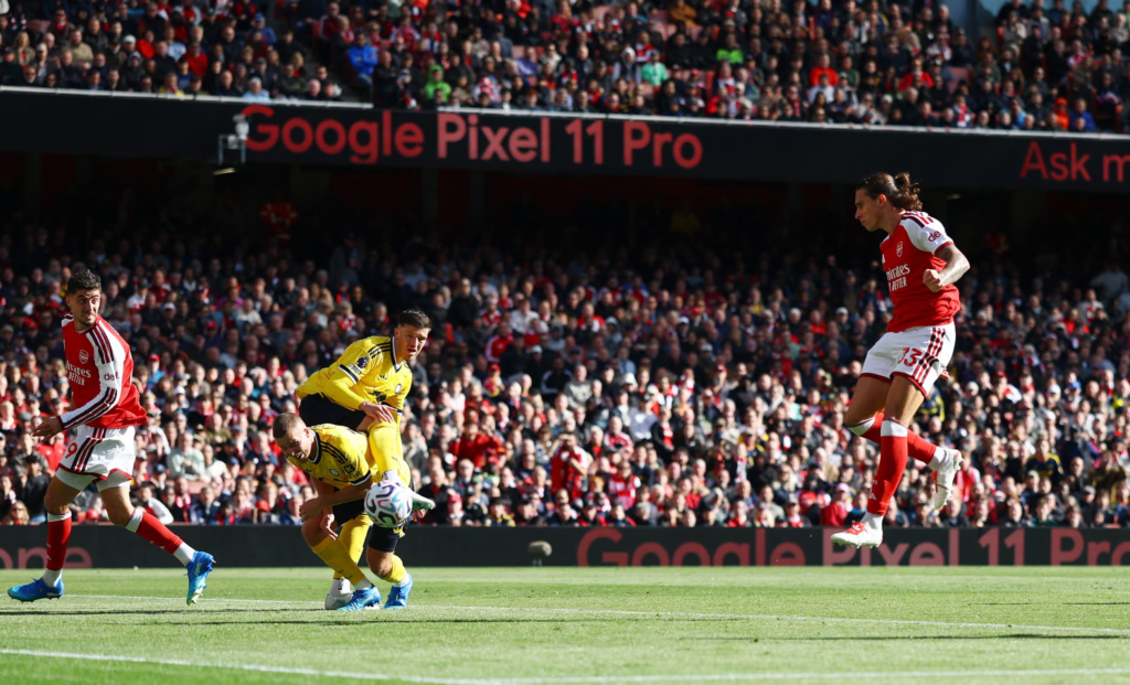 Arsenal defender Riccardo Calafiori, wearing No. 33, leaping to strike a volley in the air on the right of the frame. Two Leeds players in yellow stoop near the ball, and an Arsenal teammate in No. 9 runs in on the left, in front of a packed Emirates crowd.