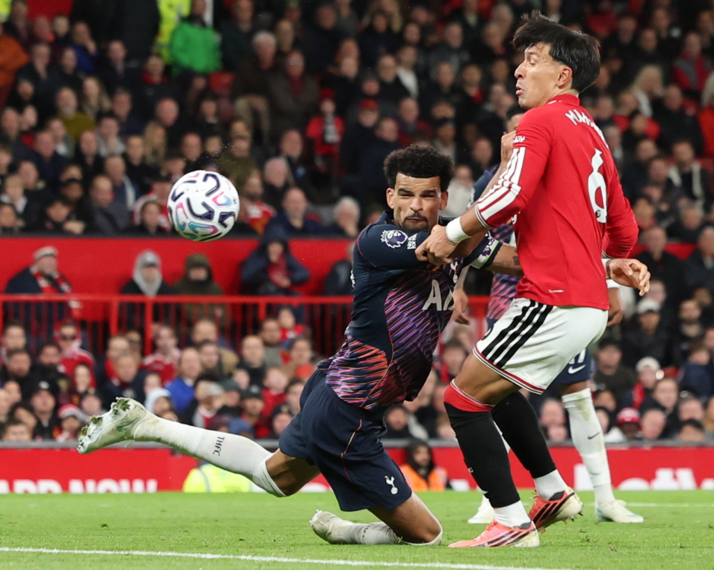 Tottenham’s Dominic Solanke, in a dark navy and purple away kit, stretched out low to the ground as he heads the ball in towards goal, with Manchester United’s Lisandro Martinez, wearing No. 6, gripping his arm beside him in front of a packed stand.