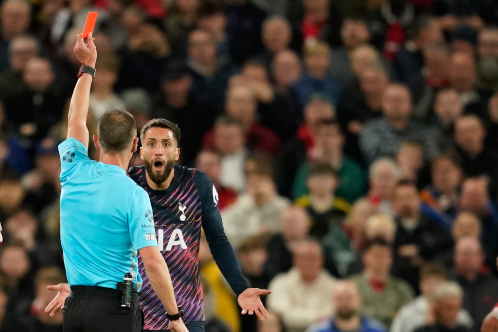 A referee in light blue with his back to the camera holding up a red card as Tottenham’s Rodrigo Bentancur, in a navy and purple kit, stares back at him, mouth open and palms upturned in disbelief, in front of a blurred crowd.