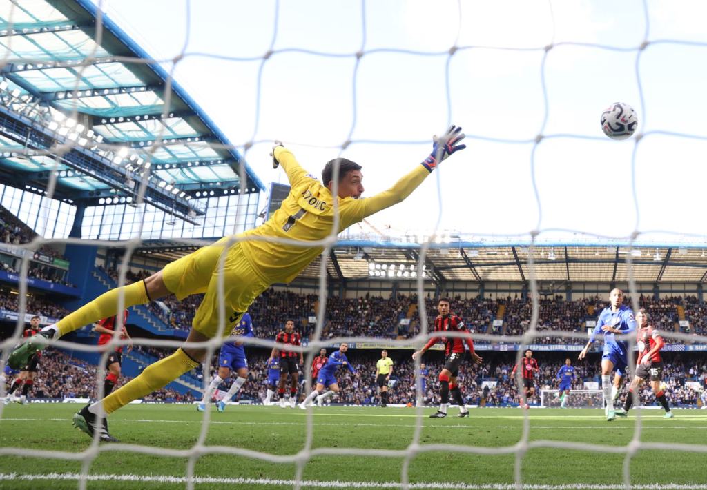 Seen from behind the goal net, Bournemouth goalkeeper Djordje Petrovic, in yellow, dives full stretch but cannot reach the ball as it flies into the goal. Chelsea and Bournemouth players look on across the pitch, with Stamford Bridge’s stands behind them.