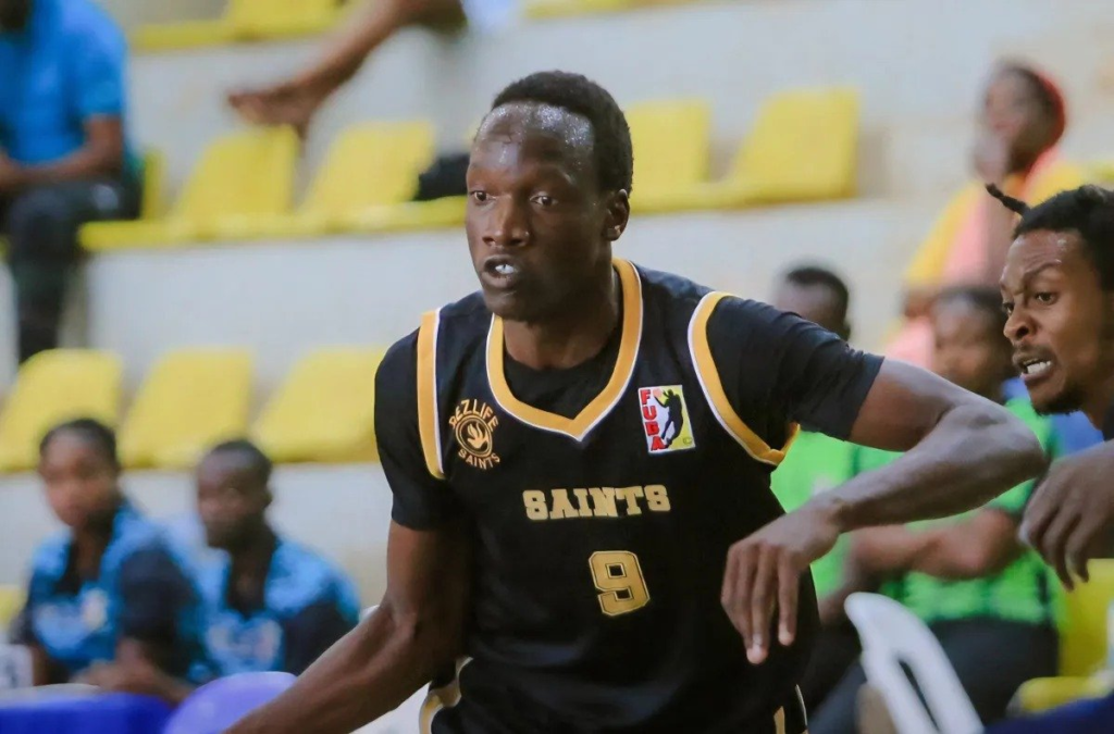 Edrine Ekau, wearing a black No. 9 Dezlife Saints jersey, dribbling during a match, with an opponent in the foreground and spectators on yellow seats behind him.
