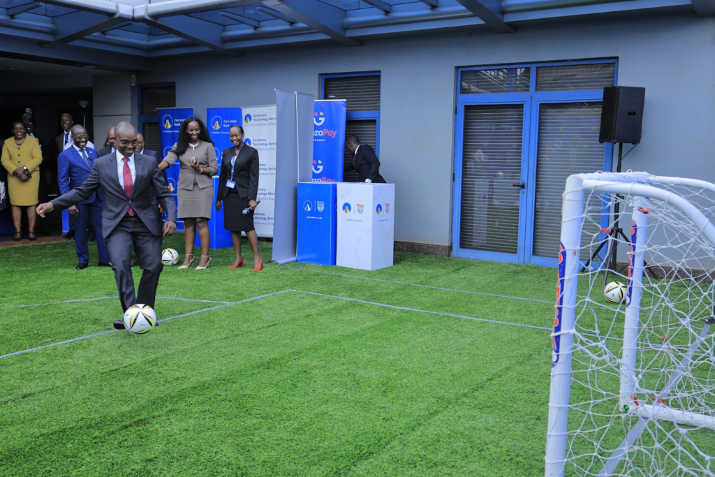 A man in a grey suit and red tie, named as Dr. Arinaitwe Rugyendo, kicks a football towards a small goal on a patch of artificial turf. Smiling onlookers in formal wear stand behind him, with Centenary Bank, Centenary Technology Services and GazaPay banners and “Partnership Launch” boxes in the background.