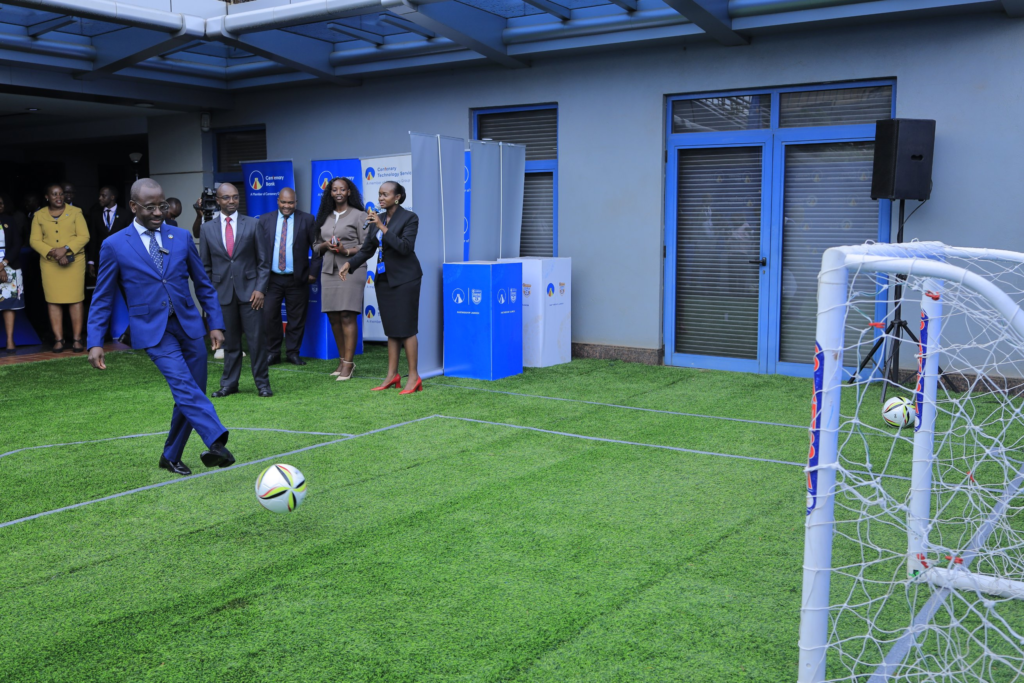 A man in a blue suit, named as Centenary Bank Managing Director Godfrey Byekwaso, kicks a football towards a small goal on a patch of artificial turf. Smiling onlookers in formal wear stand behind him, with a woman holding a microphone, Centenary Bank and Centenary Technology Services banners and “Partnership Launch” boxes in the background.
