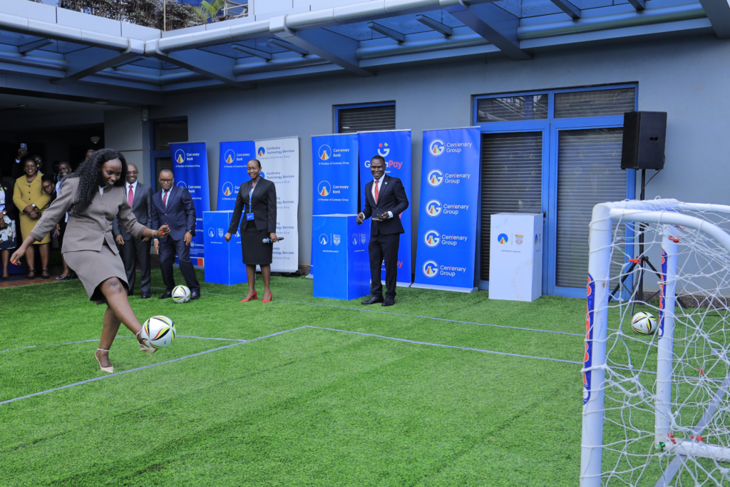A woman with long dark hair, in a brown skirt suit and beige heels, kicks a football towards a small goal on a patch of artificial turf. A woman holding a microphone, a man in a dark suit and red tie, and other smiling onlookers stand behind her, in front of Centenary Bank, Centenary Technology Services, GazaPay and Centenary Group banners and “Partnership Launch” boxes.
