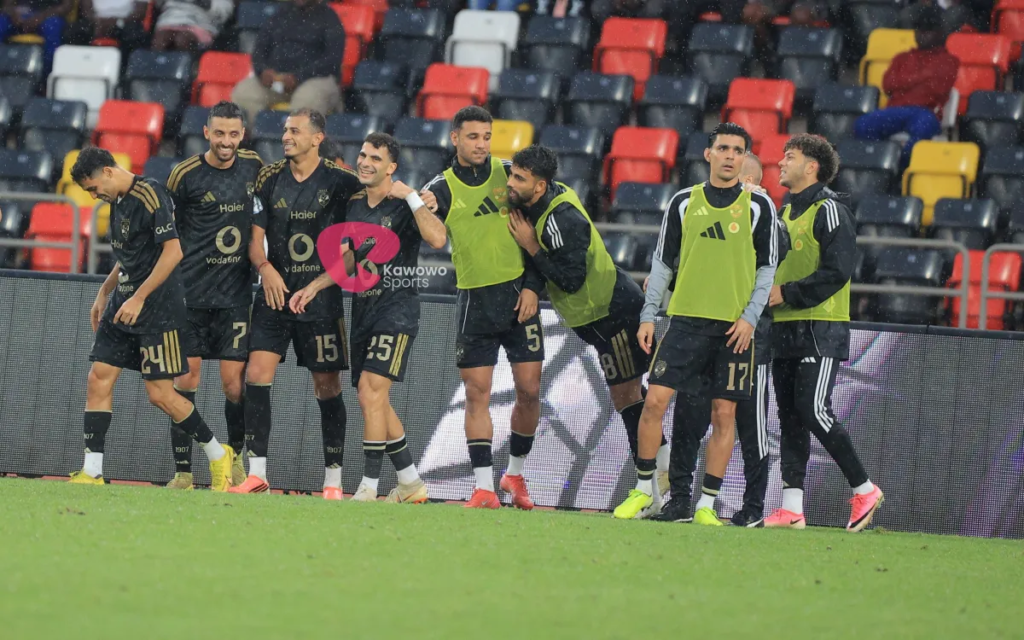 Al Ahly players in black kits and substitutes in green bibs standing in a line along the touchline, smiling and putting their arms around each other, with rows of empty red, yellow and dark seats behind them.